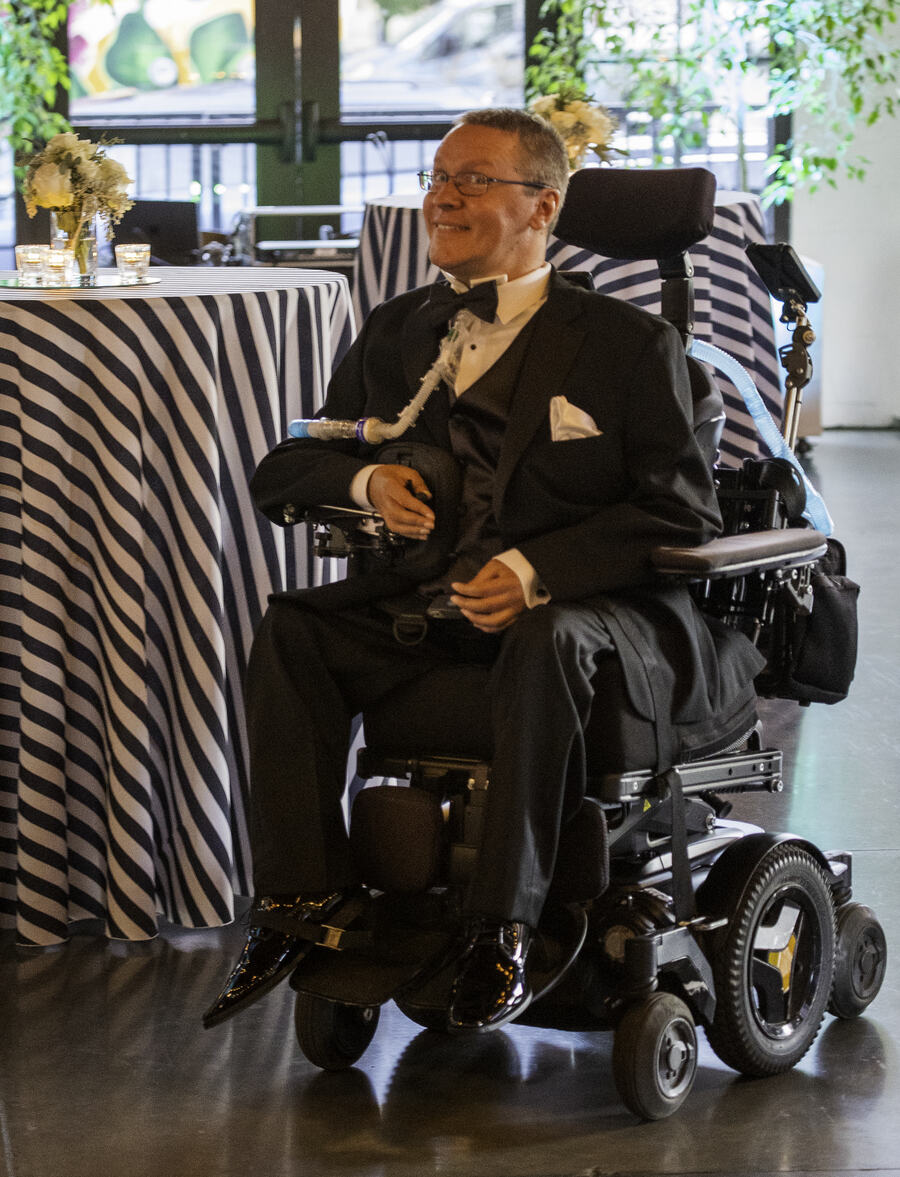 A man in a black tuxedo and bow tie, seated in a power wheelchair, smiling beside a striped cocktail table set with flowers.