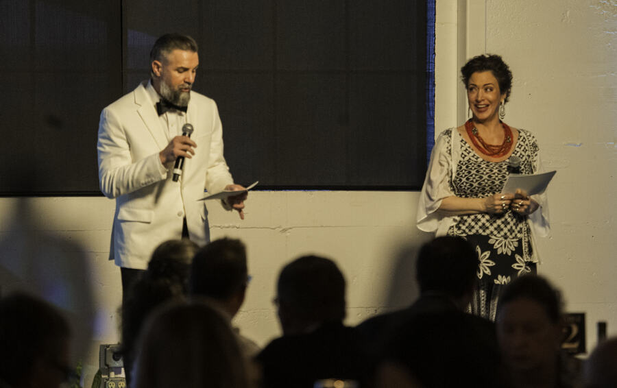 A host in a white dinner jacket speaking into a microphone while a woman beside him holds her notes, both facing the seated room.