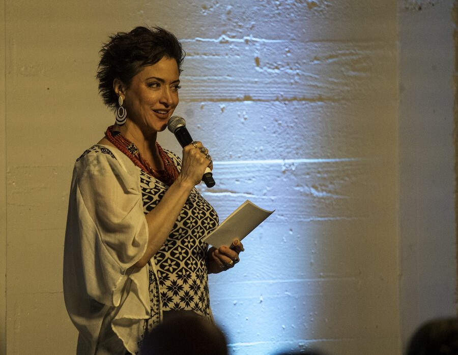 A woman in a patterned jacket speaking into a handheld microphone, holding her notes, lit against a white brick wall.