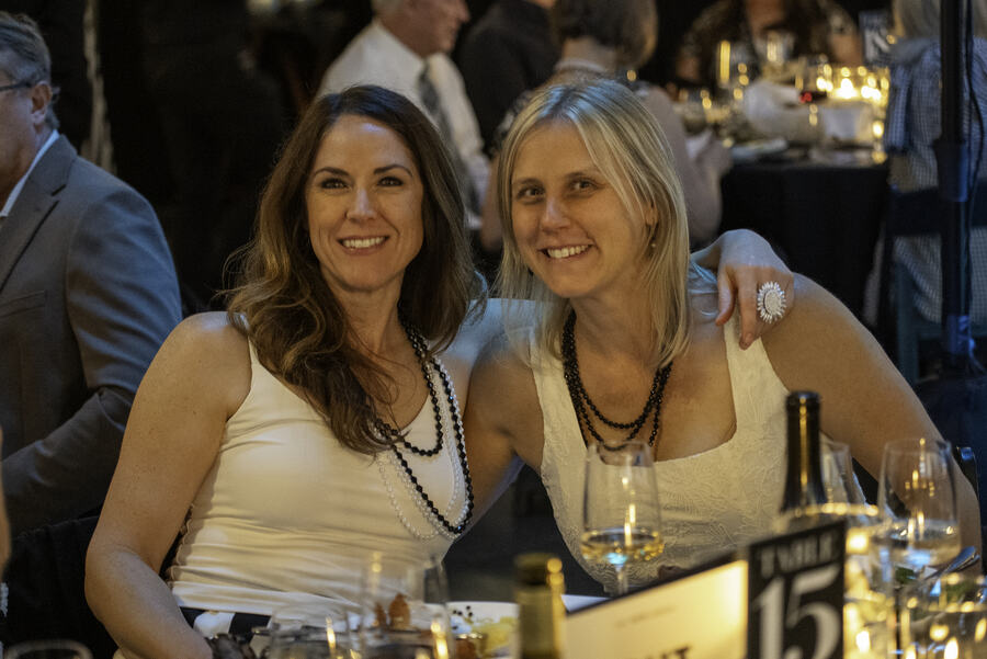 Two women in pearls leaning together at a dinner table, smiling, wine glasses in front of them.