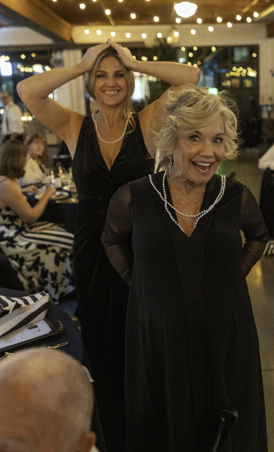 Two women in black evening dresses laughing with their hands raised to their heads during a game.