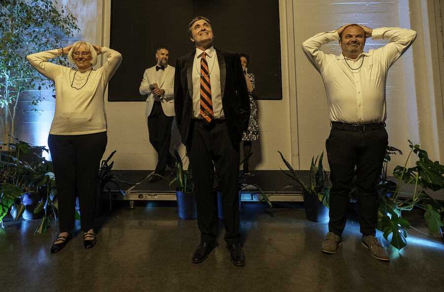 Three guests standing with their hands behind their heads mid-game, a host in a white jacket watching from behind.