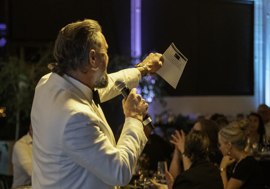 An auctioneer in a white dinner jacket holding a card aloft and calling to the room as guests bid.